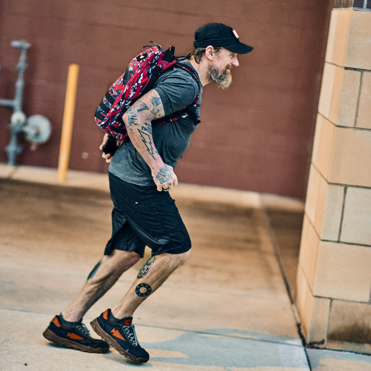 A bearded man with tattoos and a patterned backpack jogs outdoors wearing a black baseball cap, a grey t-shirt, technical cargo shorts, and athletic shoes.