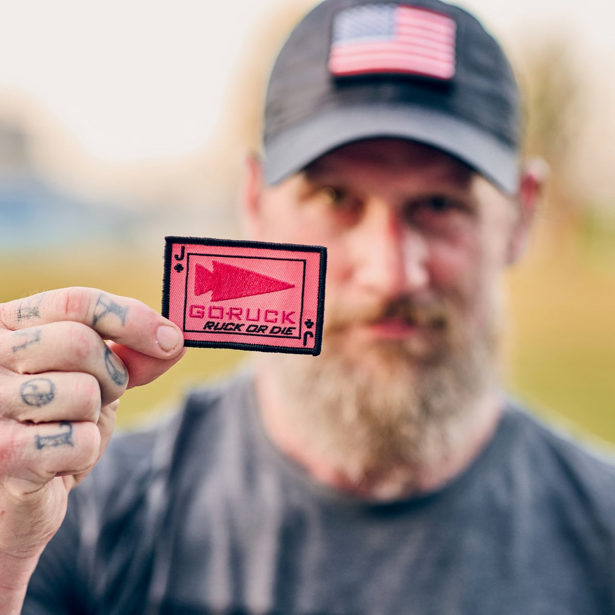 A person wearing a tactical cap with an American flag patch holds up a rectangular pink RUCK OR DIE morale patch with a spearhead icon at an endurance challenge.