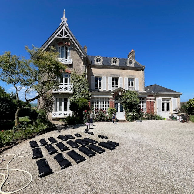 Large house with pointed roof and gravel driveway, featuring Simple Training Sandbags neatly arranged in rows—ideal for outdoor home gyms or sandbag training sessions.