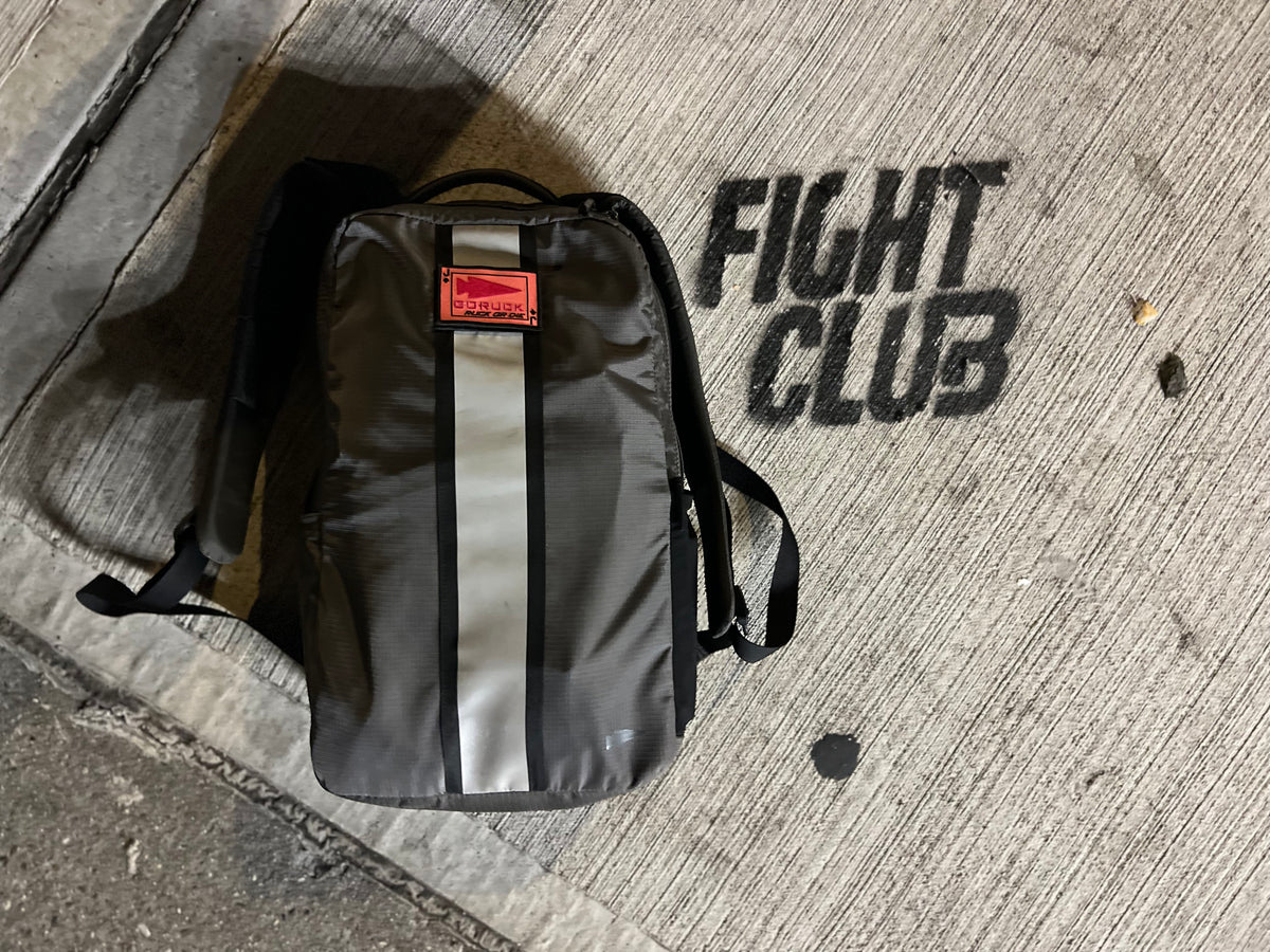 A grey GORUCK backpack with a reflective stripe and a red brand patch sits on concrete next to the words “FIGHT CLUB” spray-painted on the ground.