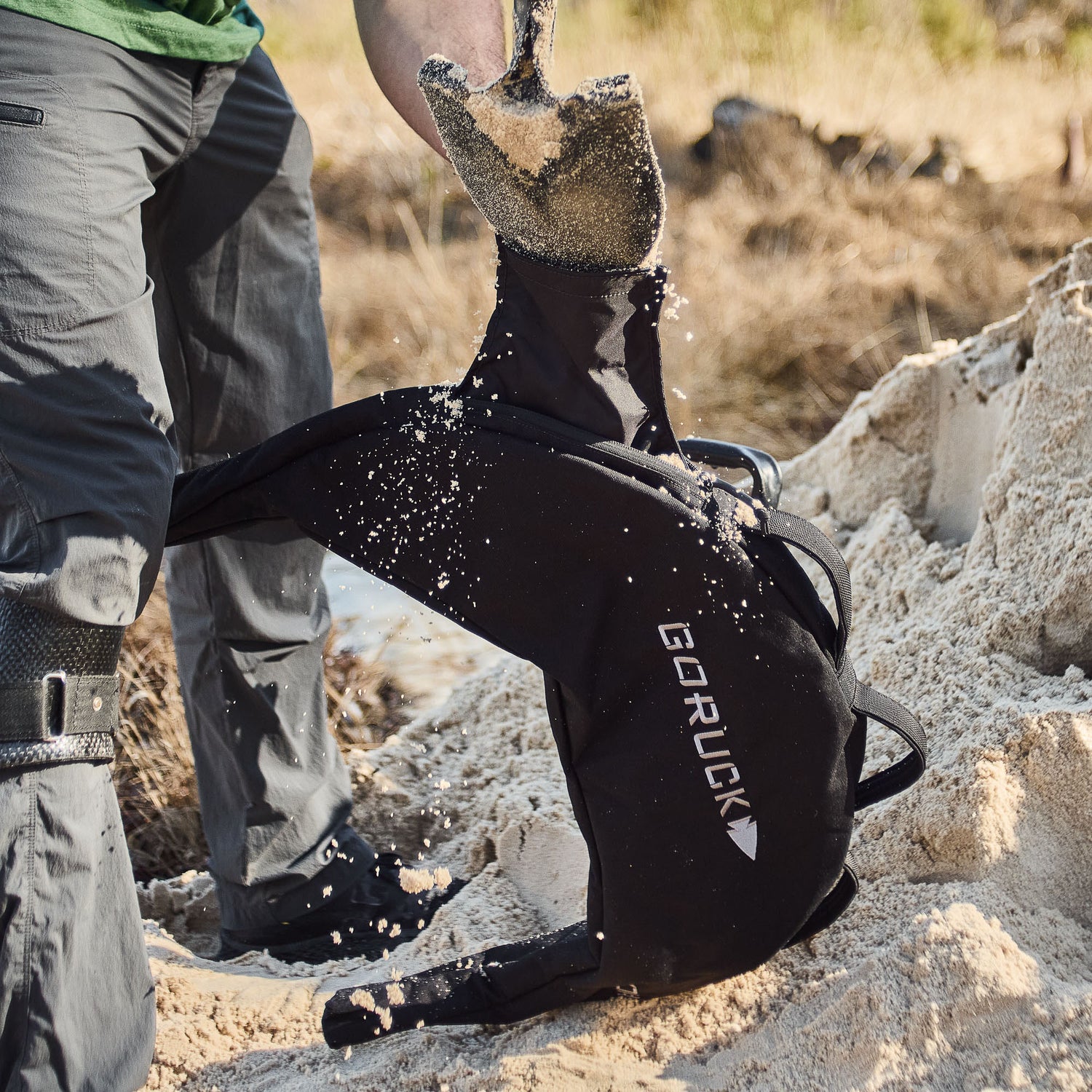 Person shoveling sand into a Bulgarian Sandbag with reinforced handles outdoors, as sand falls onto the bag.