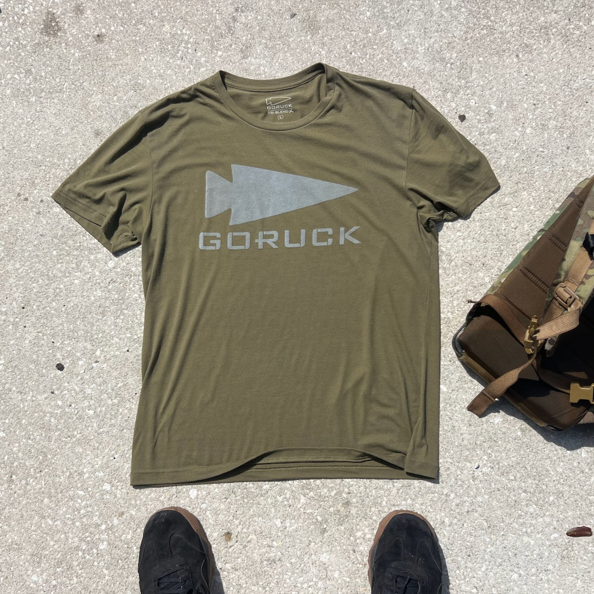 Olive green GORUCK Reflective Tee - Tri-Blend on concrete beside a bag, with black shoes in frame—highlighting Special Forces-inspired durability and style.