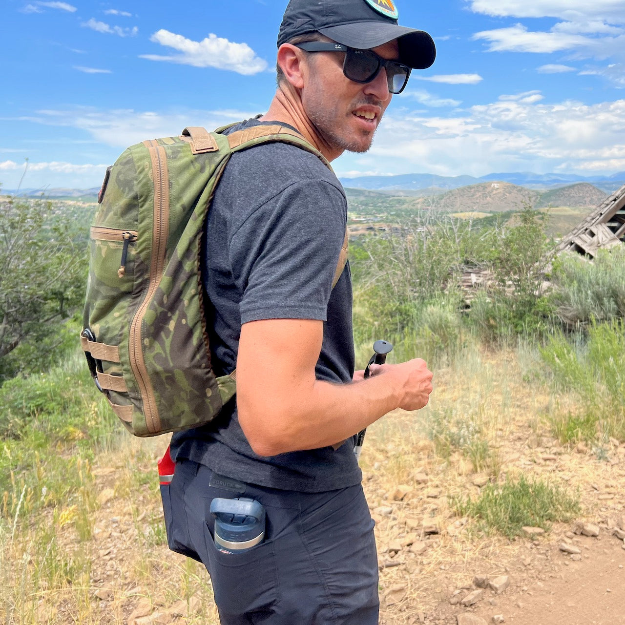 A man hikes a rocky trail under a blue sky and mountains, wearing sunglasses, a green backpack, and Men’s Simple Shorts - Lightweight ToughDry®.