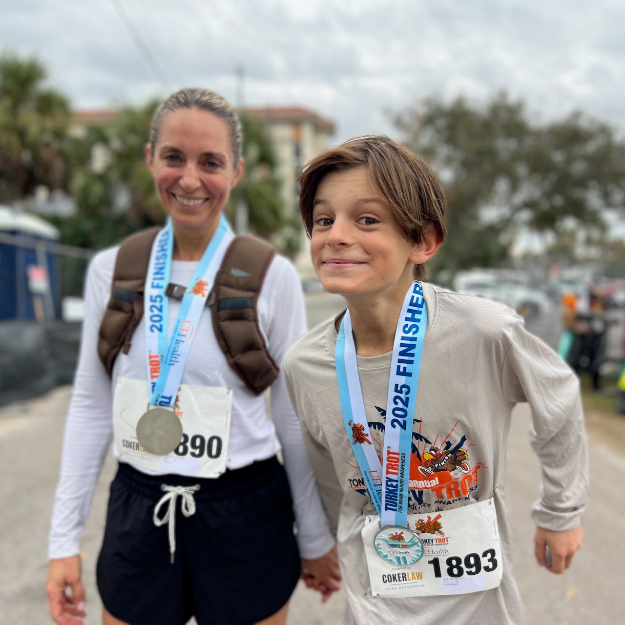 A smiling woman and boy, both wearing race medals and numbers, celebrate outdoors after finishing a 2025 event. The woman is wearing the Spy Ruck | Women's Weighted Vest.