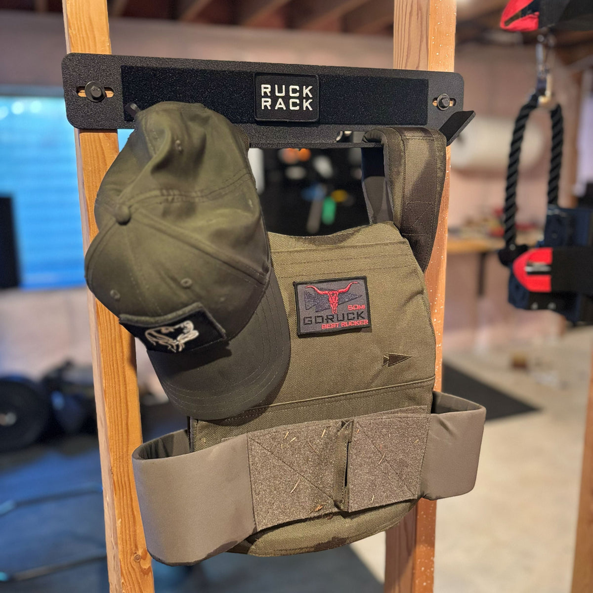 An olive green tactical weight vest and matching baseball cap hanging on a wall-mounted Ruck Rack for tactical gear organization.