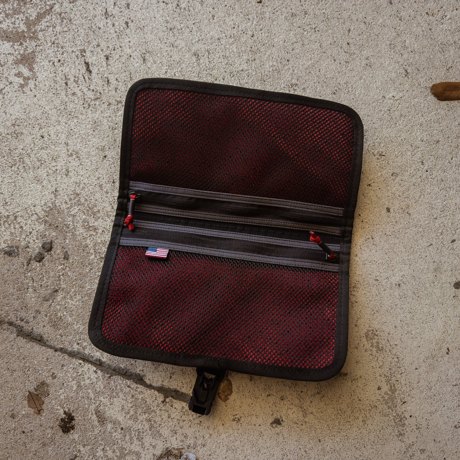 A Scars Wire Dopp pouch, made of waxed twill in red and black with mesh pockets, is open on a concrete surface.