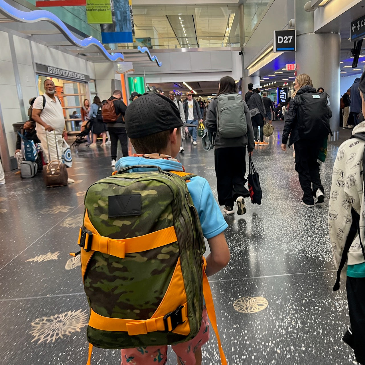 A child with a Double Bullet Ruck 18L + Bombproof Laptop Compartment camo backpack walks through a busy airport terminal, travelers ahead, ready for adventure.