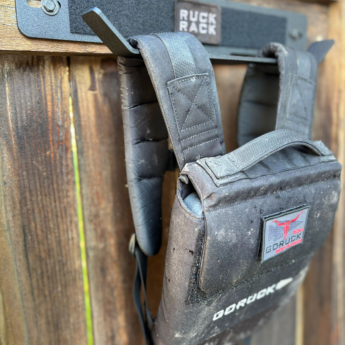 Black GORUCK rucksack hanging on a metal wall-mounted storage rack attached to a wooden fence for tactical gear organization.