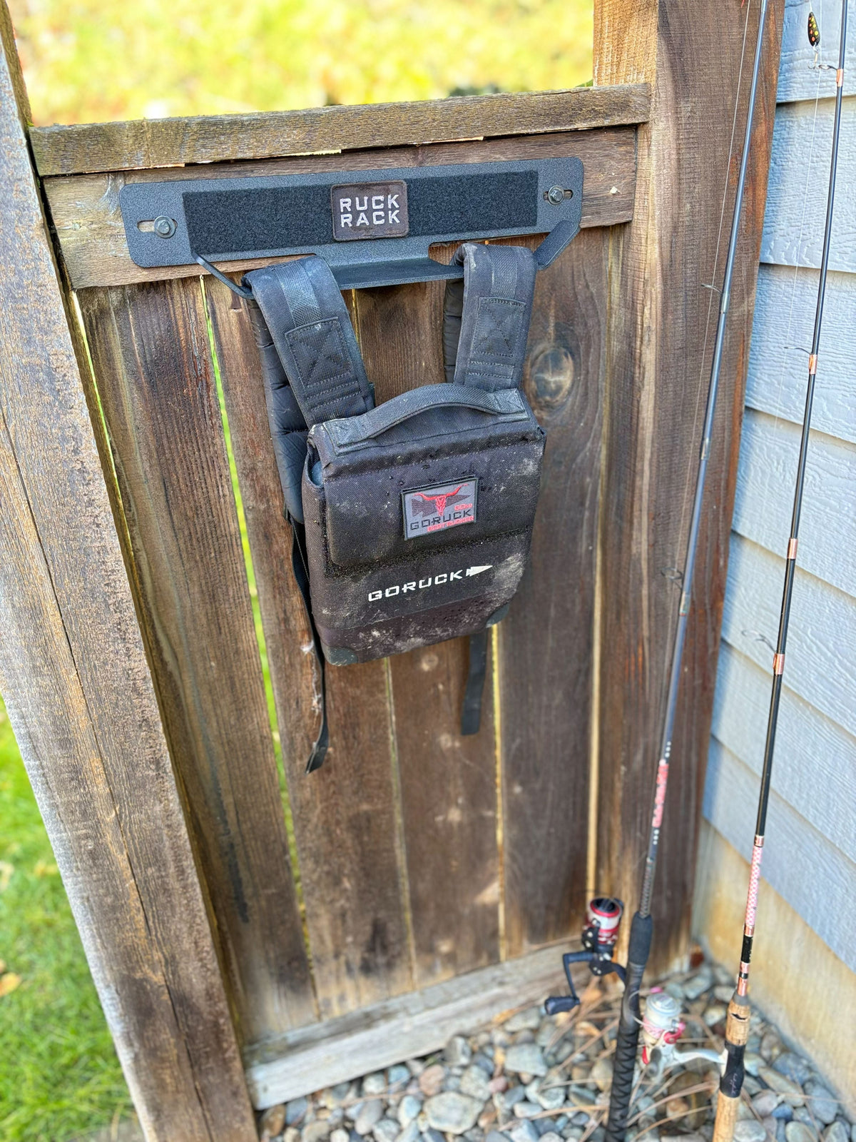 Black GORUCK rucksack hanging on a wall-mounted storage rack attached to a wooden gate, with fishing rods leaning against the side for tactical gear organization.