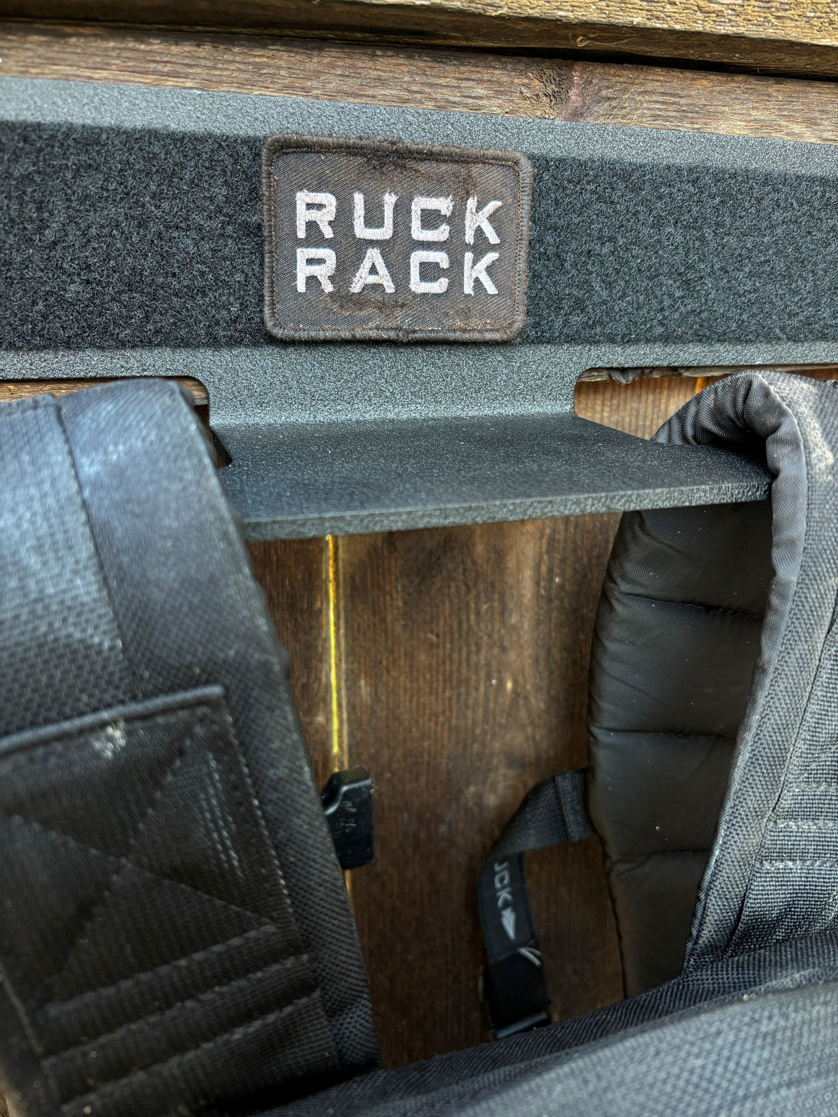 A black metal wall-mounted gear hanger with a “RUCK RACK” patch, used for tactical gear organization while holding the shoulder straps of a black backpack.