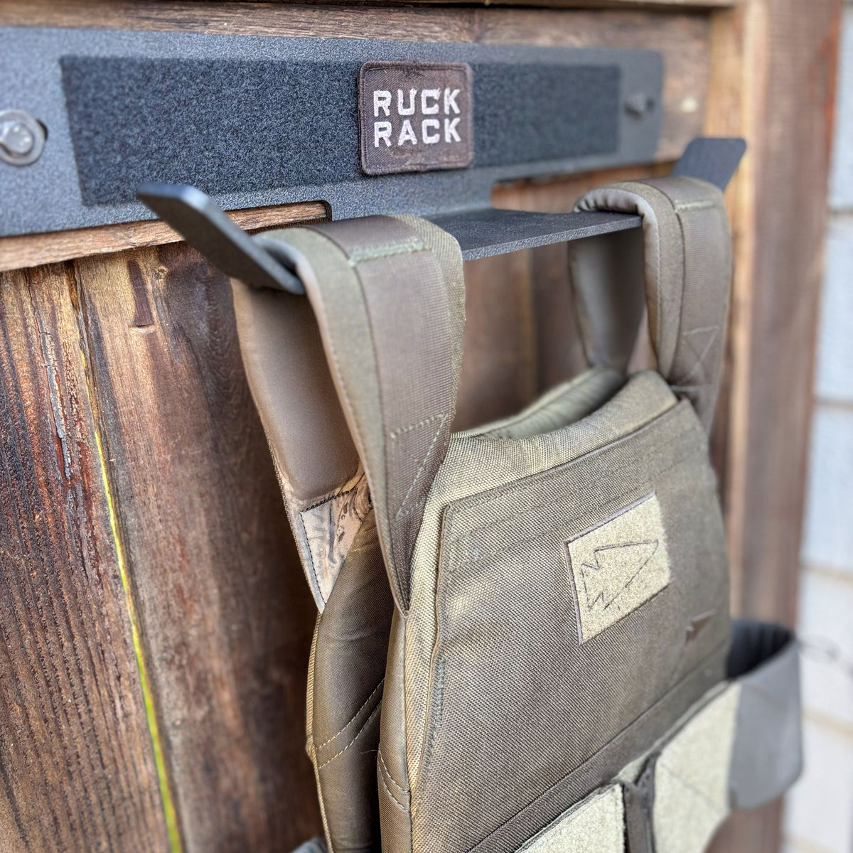 An olive green tactical weight vest hangs on a black metal wall-mounted storage rack designed for tactical gear organization.