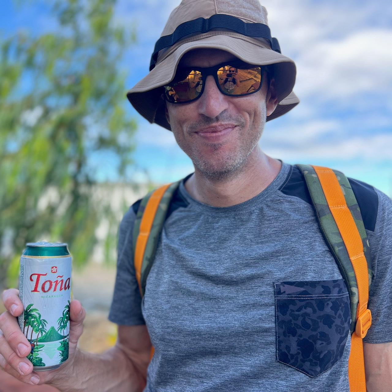 A smiling man in sunglasses and a hat holds a Toña beer, sporting a grey shirt and carrying the Double Bullet Ruck 18L with Bombproof Laptop Compartment on his back outdoors.