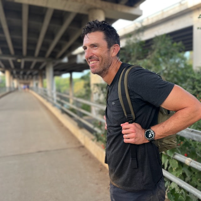 A man with a backpack stands on a pedestrian bridge beneath an overpass, smiling as he looks off into the distance with classic simplicity.
