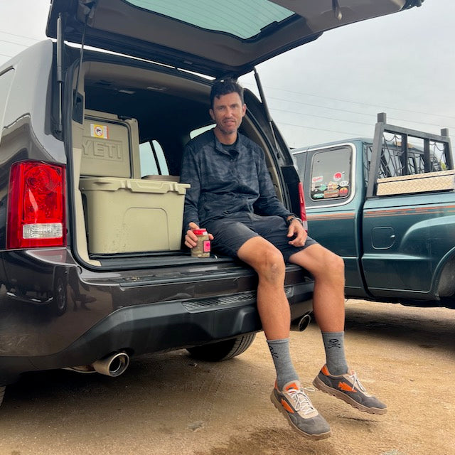 A man sits on the open tailgate of an SUV wearing an abrasion resistant shirt, holding a small jar next to two coolers.