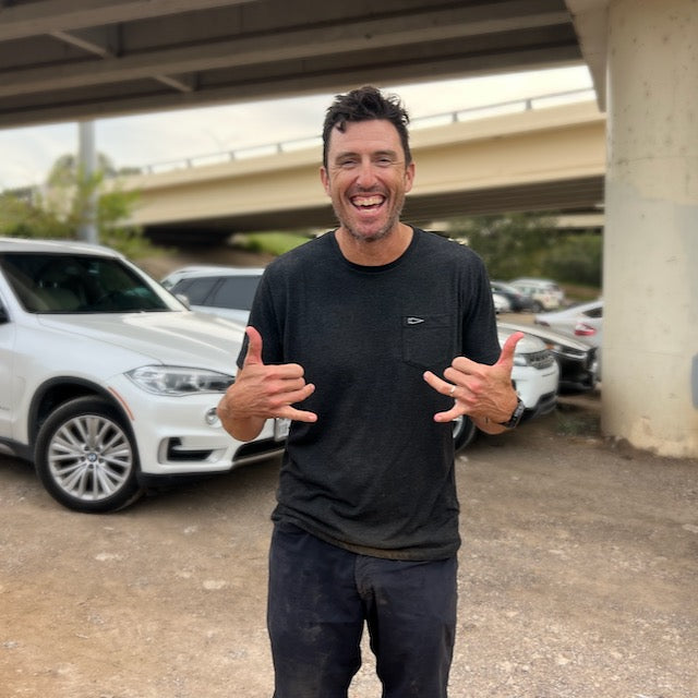 A smiling man in a dark t-shirt with classic simplicity makes the shaka hand gesture with both hands while standing in a parking lot.