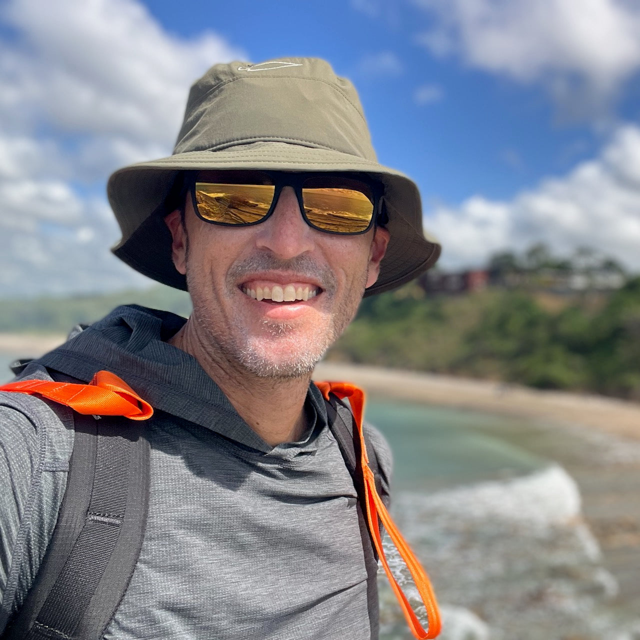 A man wearing the Boonie Hat - Slick - ToughDry and sunglasses smiles outdoors by the beach, with waves and greenery in the background.