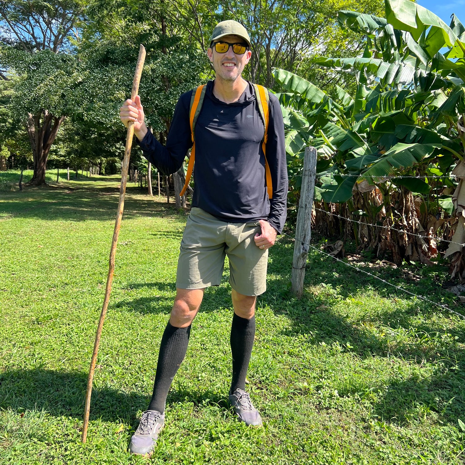 Man in outdoor gear stands on grass with a walking stick, wearing sunglasses, a backpack, and the Performance Running Hat - Italian ToughMesh made from premium Italian fabric.