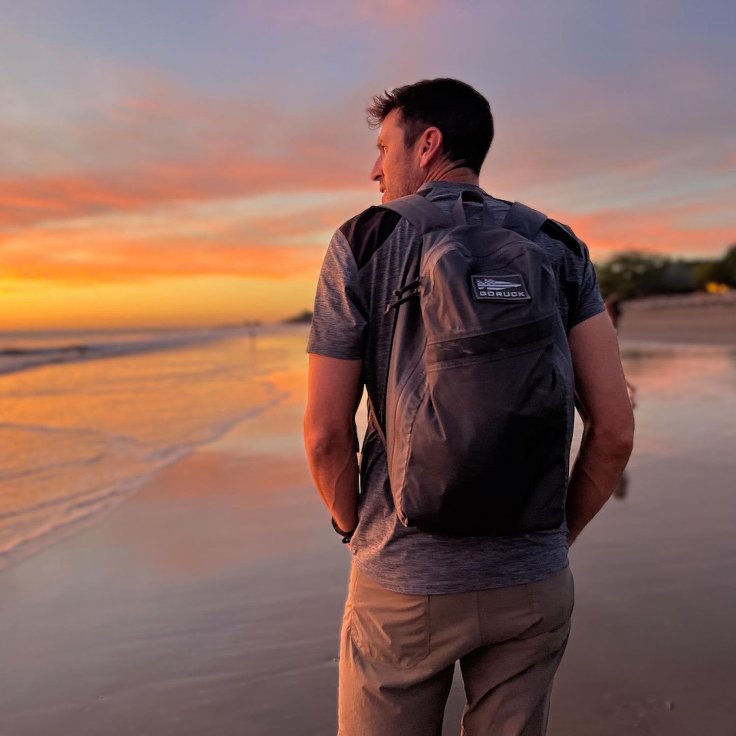 A man with the Packable Bullet Ruck - Ripstop ROBIC® backpack stands on a beach at sunset, gazing out at the ocean and colorful sky.