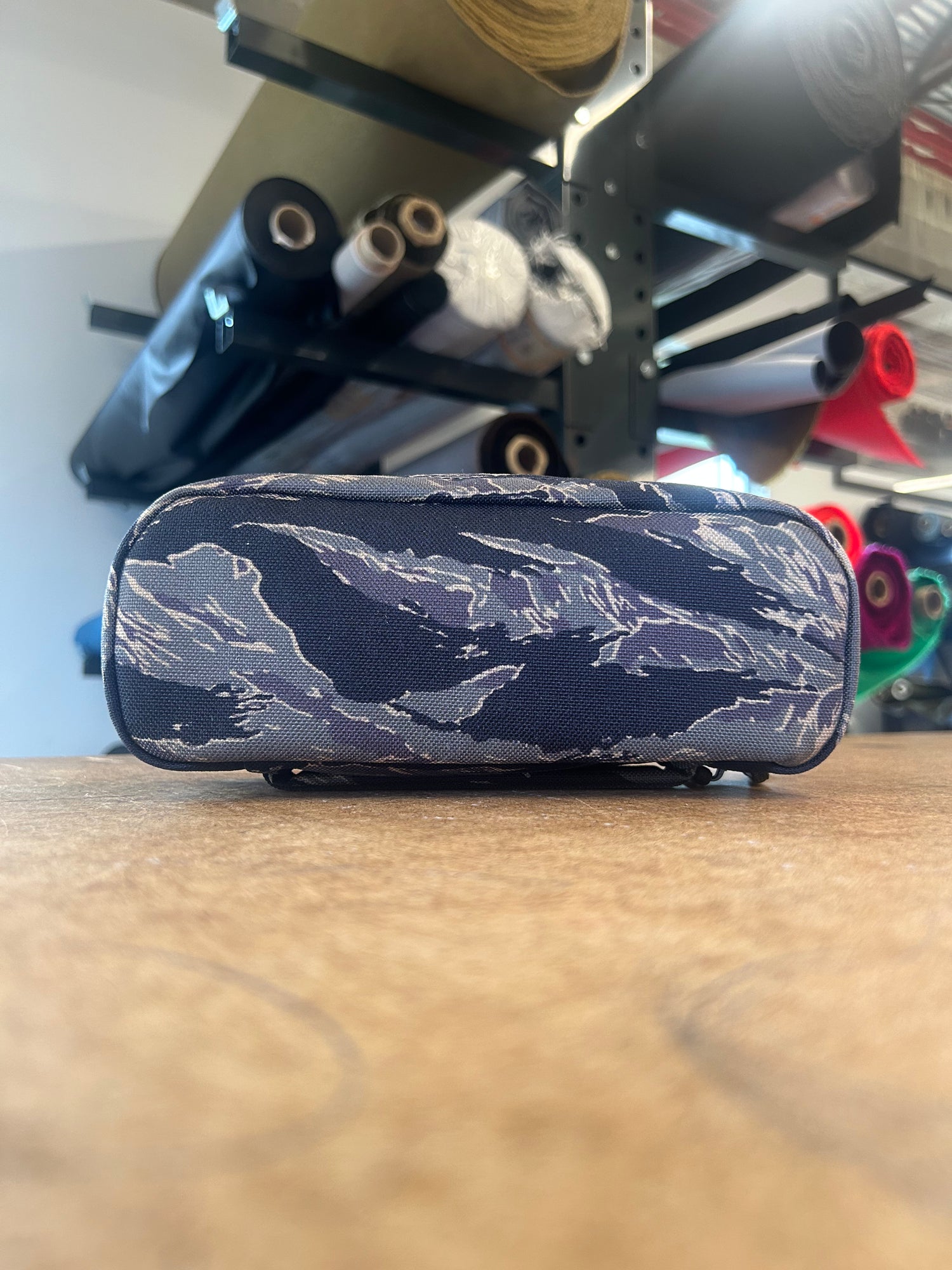 The Shadow Pocket- Tiger Stripe, a blue Cordura bag ideal for rucksack organization, rests on a wooden table with softly blurred fabric rolls in the background.