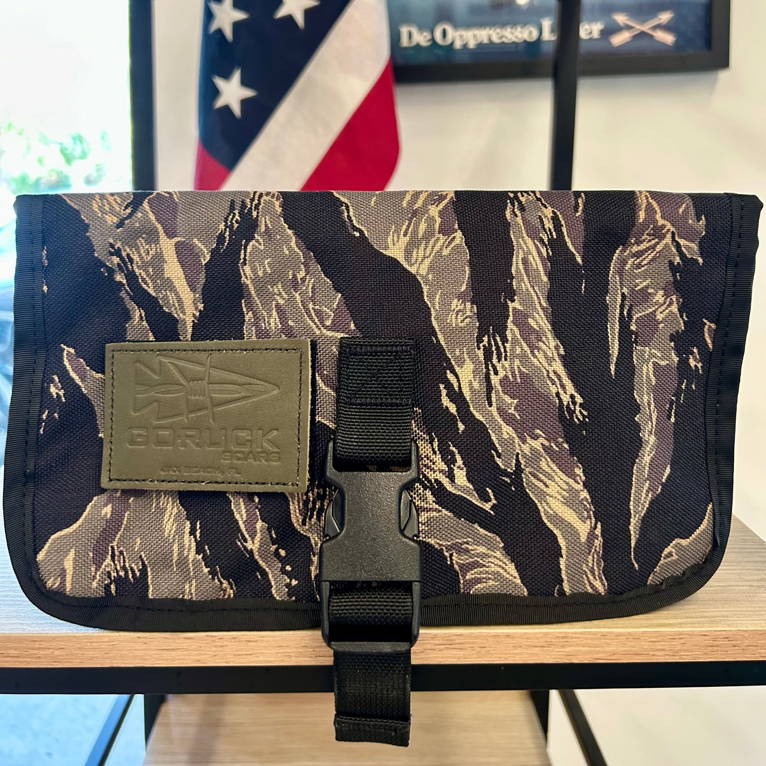 The Scars Wire Dopp is a camouflage pouch with a buckle clasp and GORUCK patch, ideal as a travel organizer or for carrying EDC essentials, shown in front of a U.S. flag.
