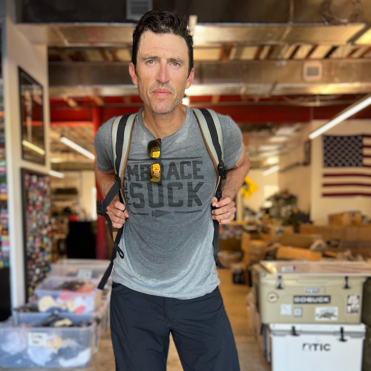 A man with a backpack stands indoors, wearing sunglasses on his collar and the gray Embrace the Suck Tee - Tri-BlendX, a nod to the USMC's official motto.