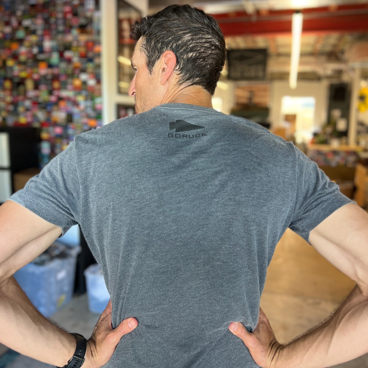 A man stands indoors with his back to the camera, hands on hips, wearing the Embrace the Suck Tee - Tri-BlendX in gray, featuring GORUCK's design and the USMC motto: