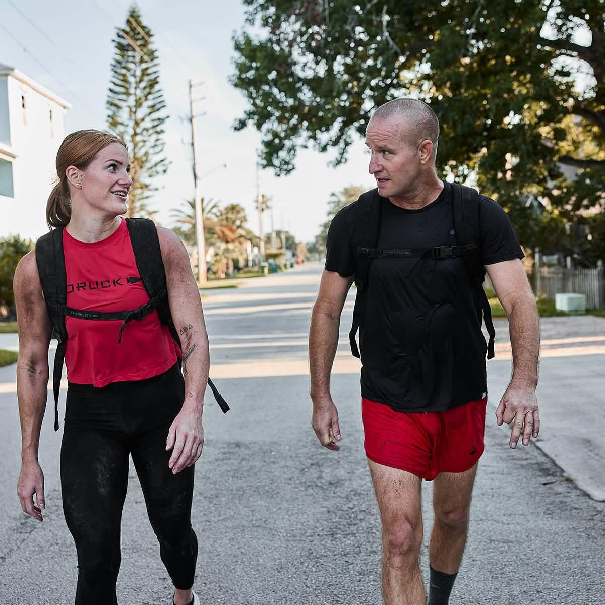 Two fit adults walking on street wearing GORUCK gear backpacks, red and black activewear during daylight