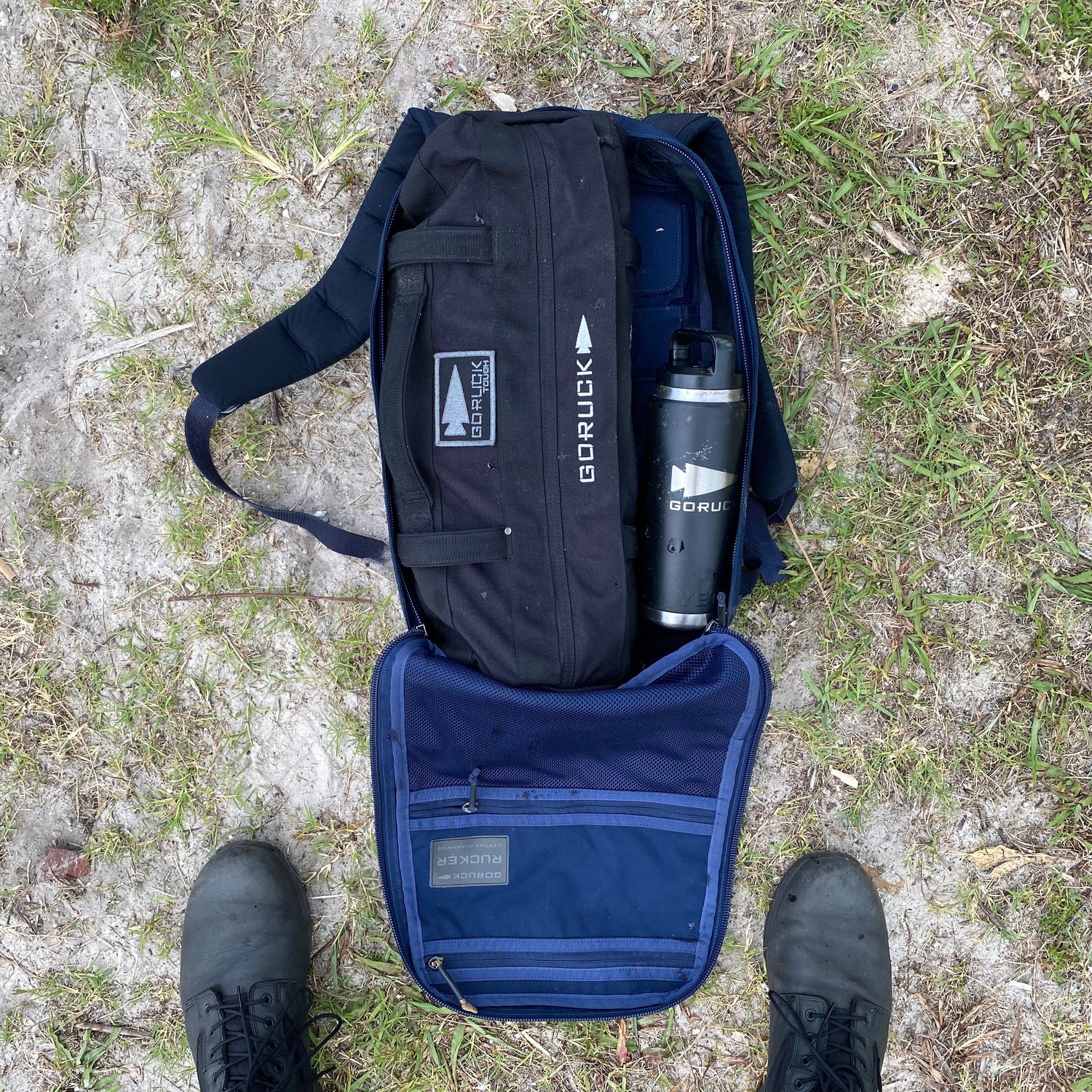 On grass, an open backpack reveals Simple Training Sandbags and a water bottle inside—ideal for sandbag workouts or your home gym. Black boots are visible below.