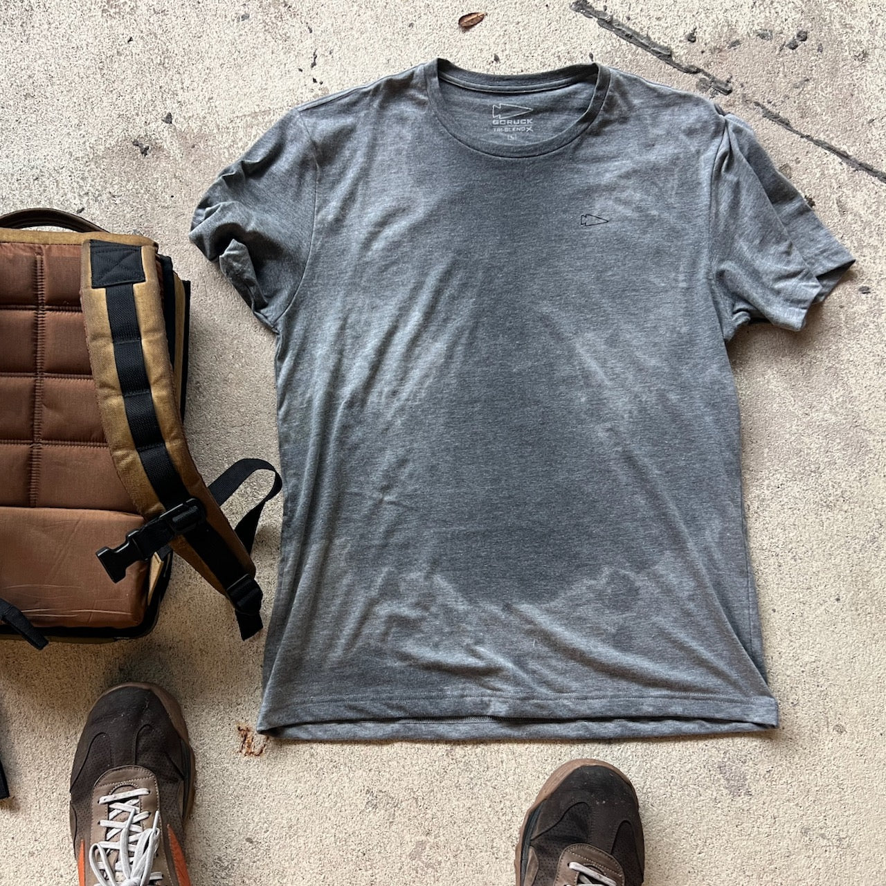 A sweat-stained gray Subtle Spearhead Tee - Unisex Tri-Blend lies on concrete beside a brown backpack and two sneakers.