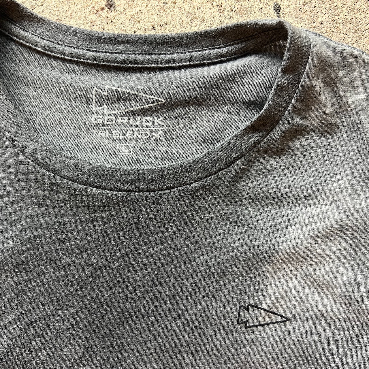 The Subtle Spearhead Tee - Unisex Tri-Blend is a gray t-shirt with a small arrow logo on the chest and inside the collar, inspired by Special Forces and featuring Anti-Stink technology.