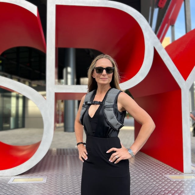 A woman stands confidently in front of a bold red and white “SPY” sign, wearing the Spy Ruck | Women's Weighted Vest and sunglasses, embodying GORUCK-inspired style.