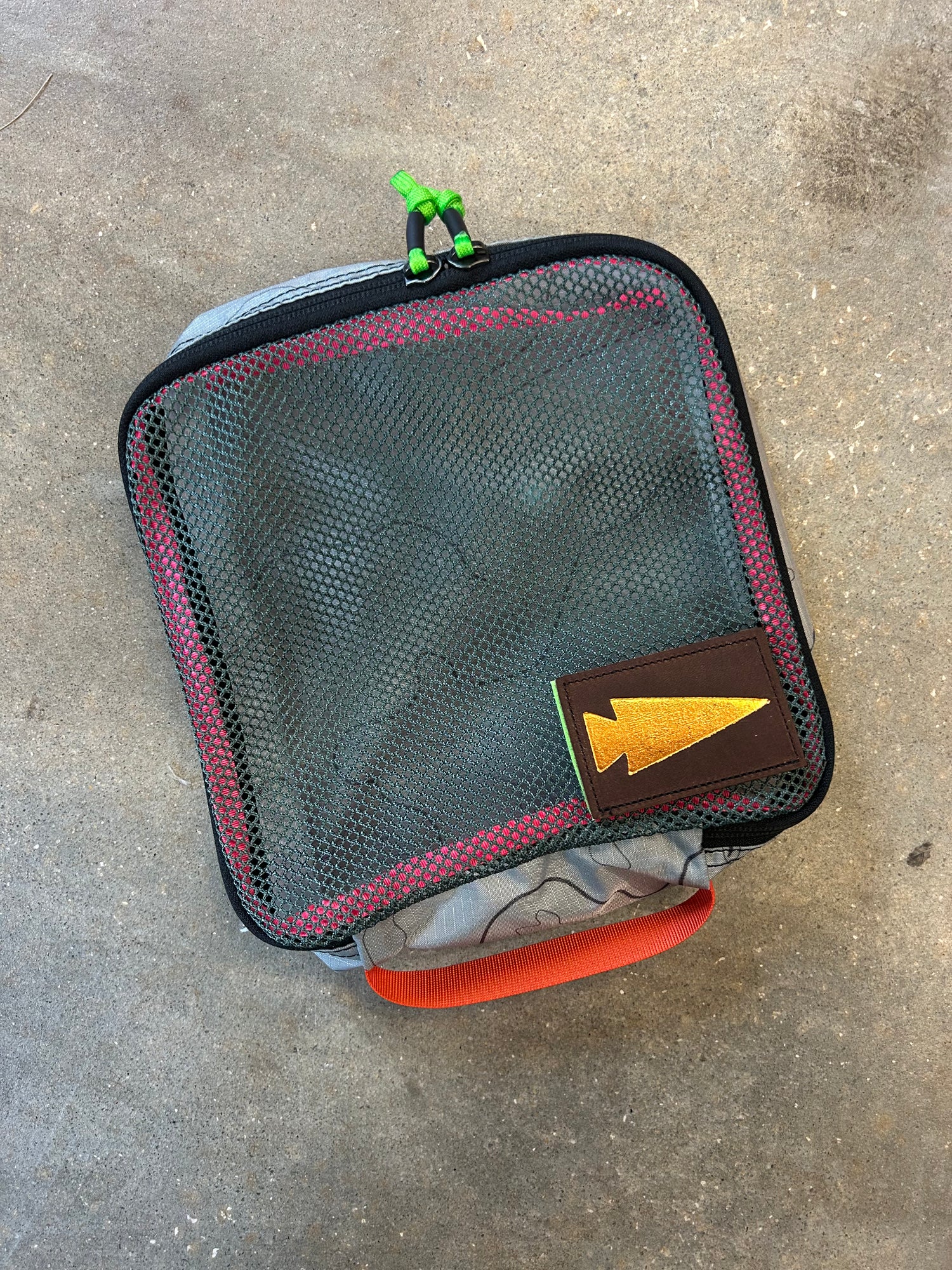 The Ripstop Robic XL Packing Cube is a square mesh pouch with a red zipper, green pull cord, orange handle, and yellow arrow patch—ideal for travel organization with your favorite packing cubes.