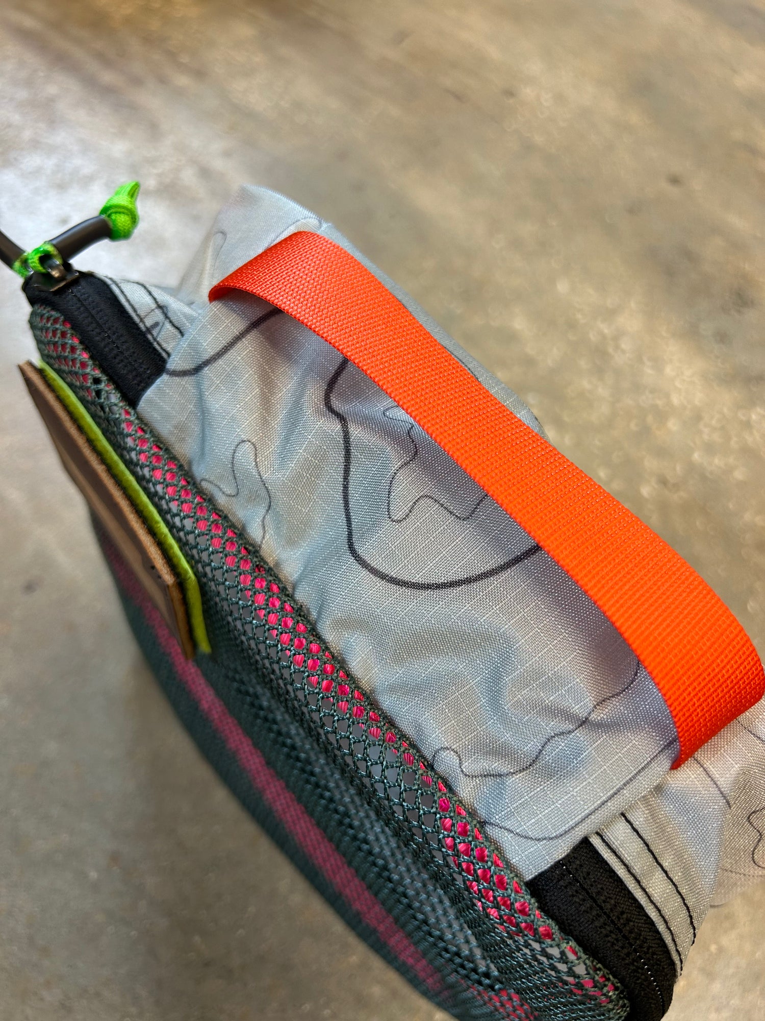 Close-up of the Ripstop Robic XL Packing Cube in gray, made from durable nylon with a bright orange handle and green zipper pull—perfect for travel organization or use with other packing cubes, shown on a textured floor.