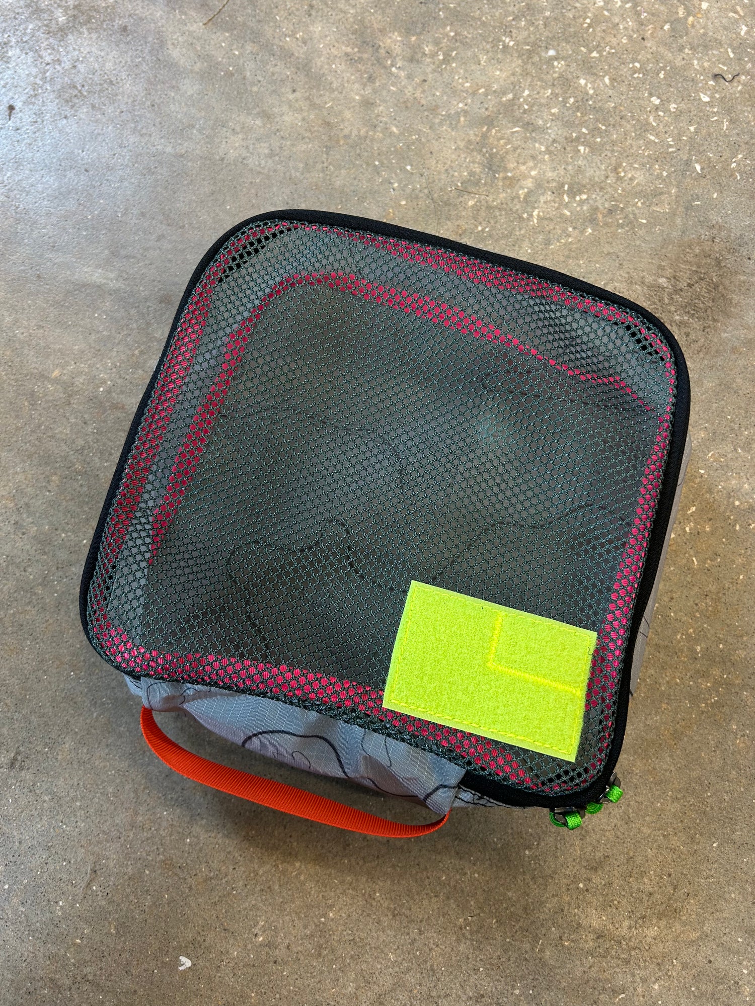 The Ripstop Robic XL Packing Cube, featuring a black mesh top, red trim, orange handle, and bright green label, rests on concrete—perfect for keeping your travel essentials organized.