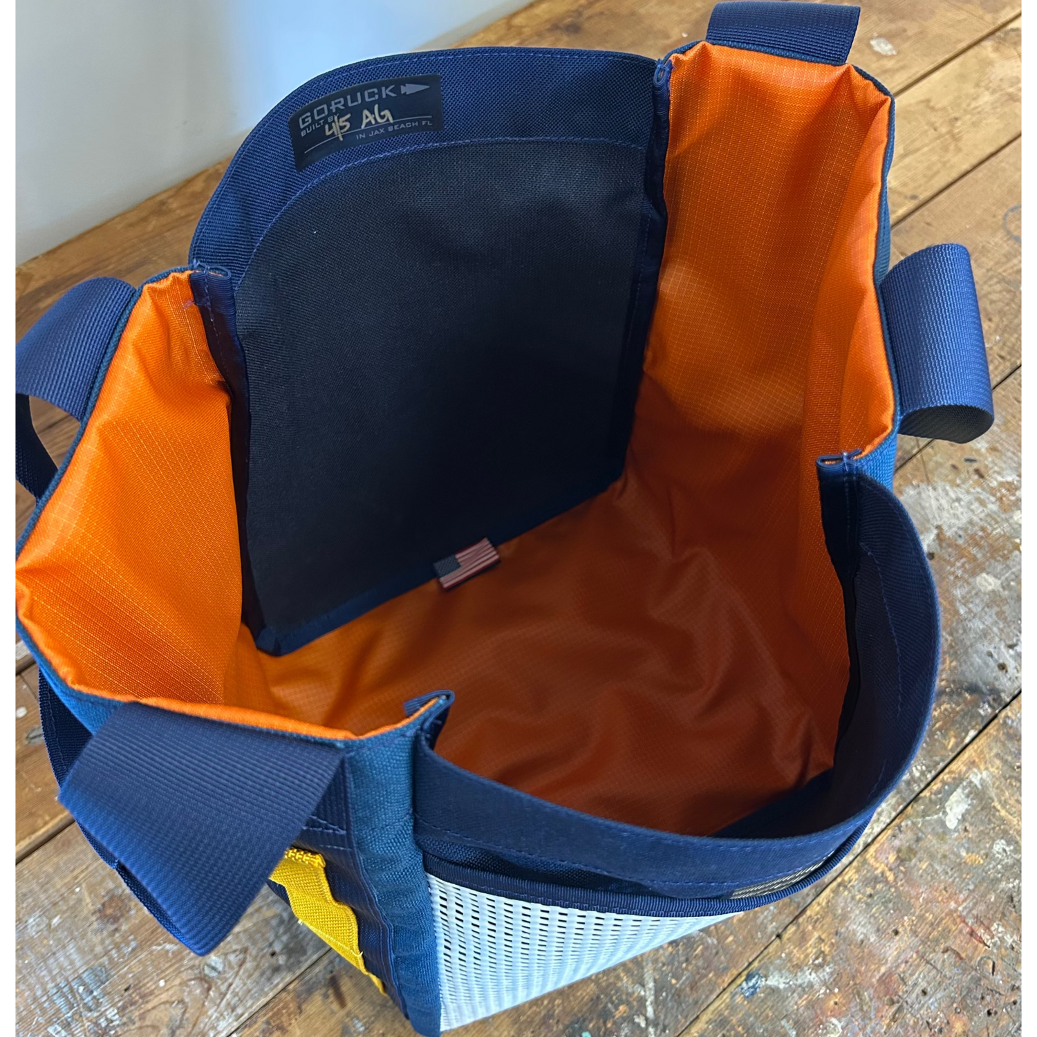 The Scars Tote, crafted from 1000D Cordura with a blue exterior, orange interior lining, and sturdy handles, sits on a wooden surface—an everyday tote bag designed for durability.