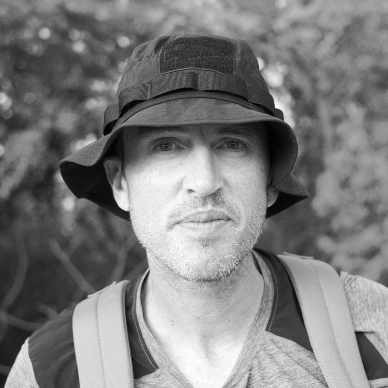 A man wearing a Tactical ToughDry Boonie Hat and backpack looks at the camera outdoors, with blurred trees in the background.