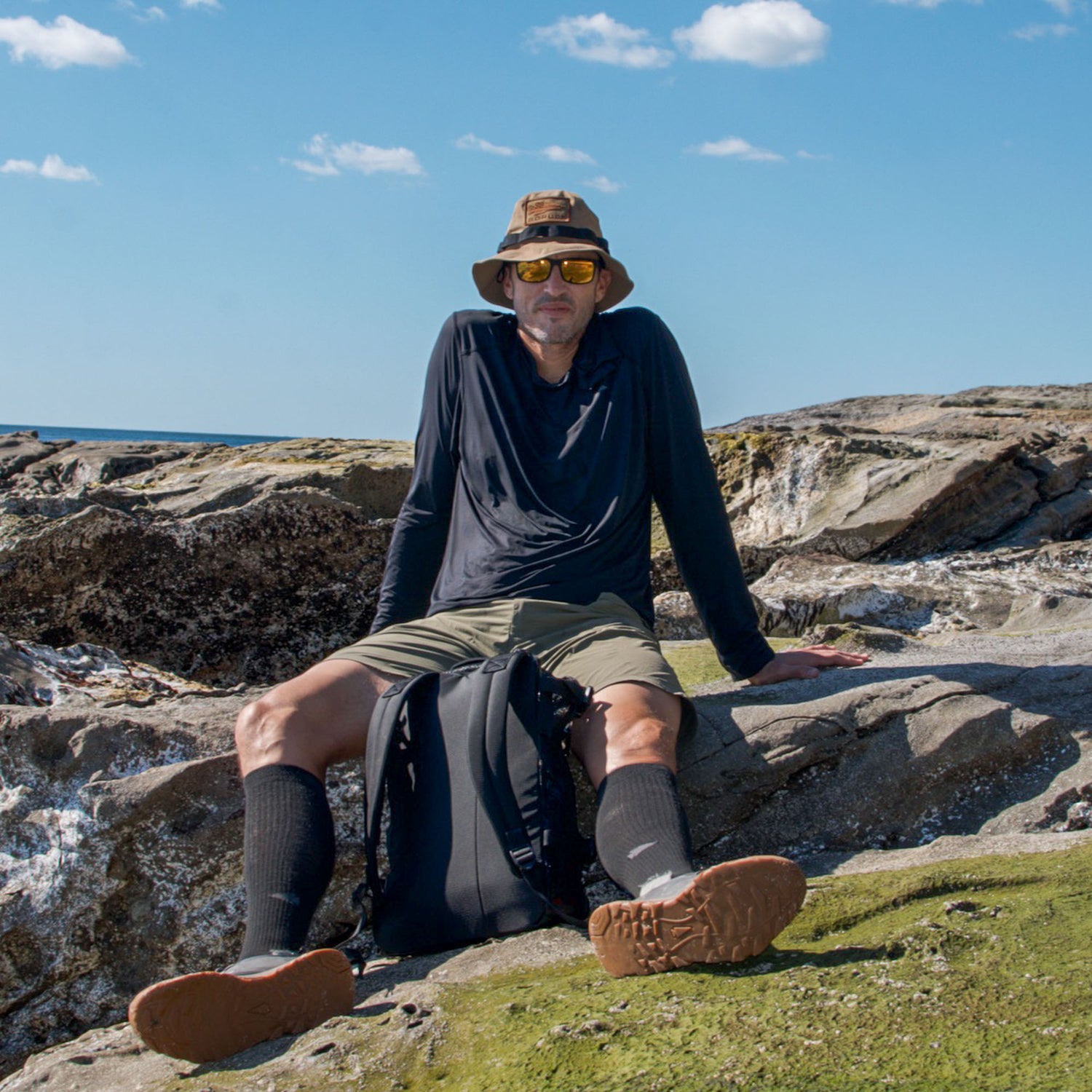 A man in hiking attire sits on rocky terrain under a blue sky with a backpack at his feet, wearing Merino Compression Socks for comfort during long treks.