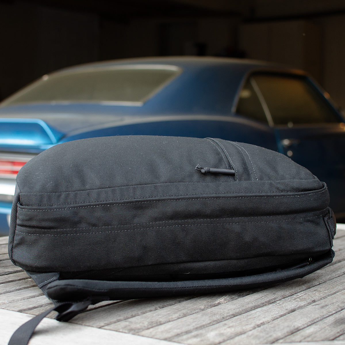 The GR0 USA Slick - Ballistic Nylon Cordura backpack in black rests on a wooden surface, with a classic blue car softly blurred in the background.