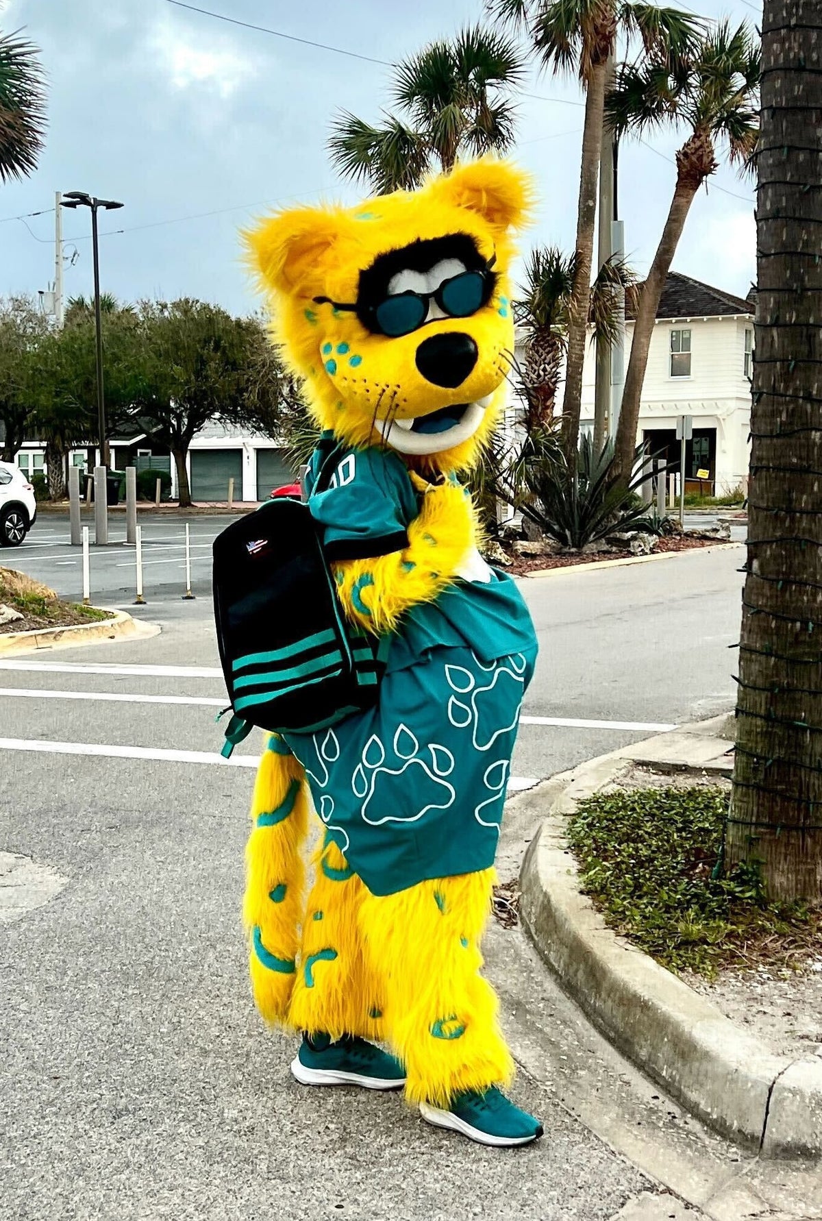 A yellow jaguar mascot wearing sunglasses, a teal jersey, and a black backpack stands on a street corner representing the Jacksonville Jaguars Foundation.