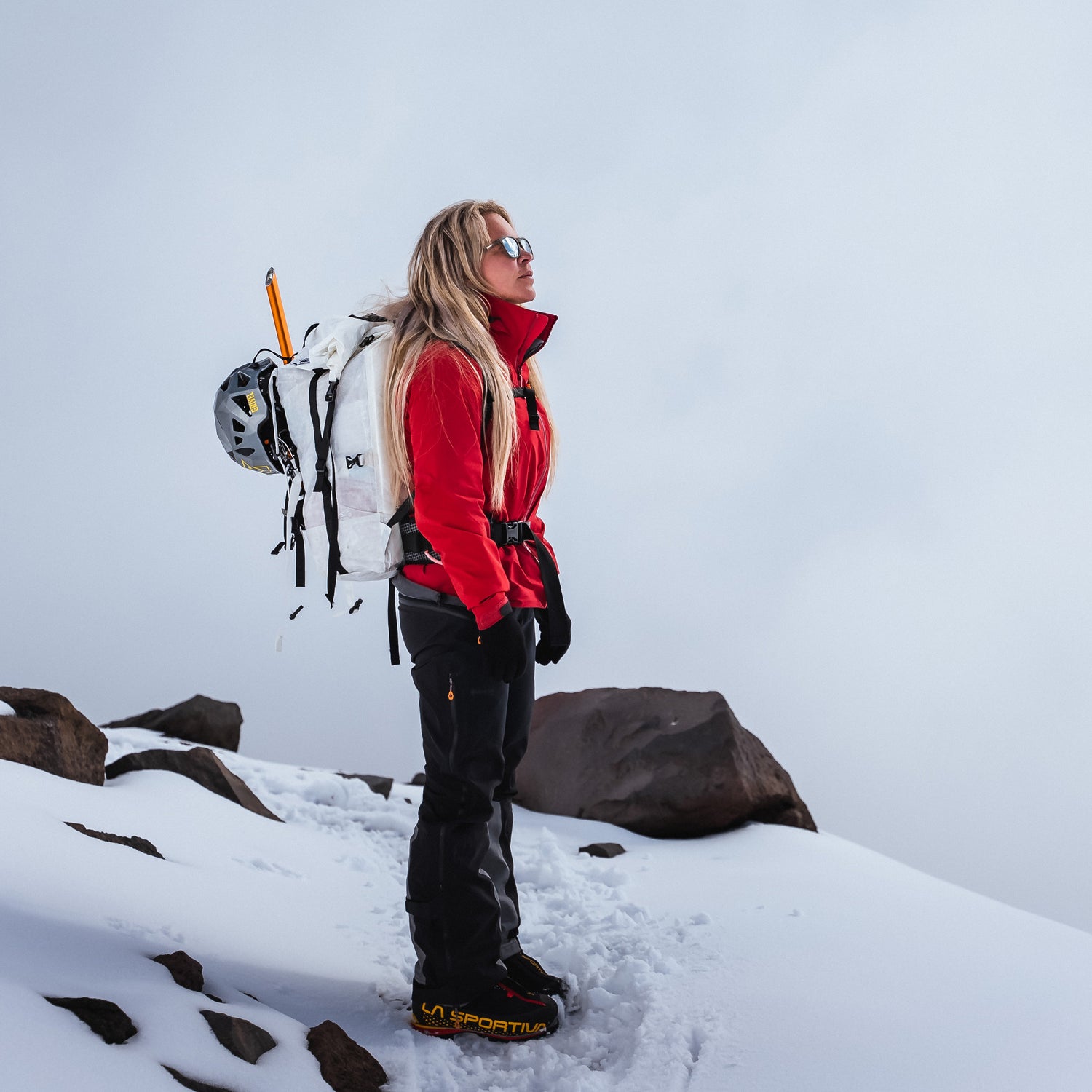 A woman in winter gear stands on a snowy mountain, looking up, with rocks and snow around her.