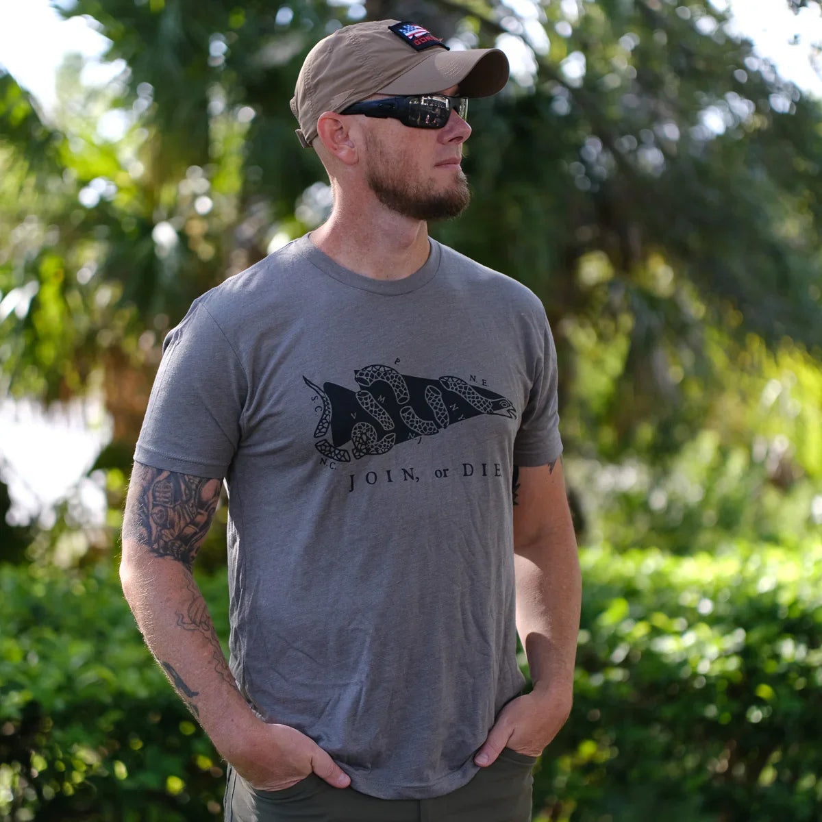 Man wearing GORUCK join or die graphic t-shirt in Venetian grey, sunglasses, and cap outdoors with greenery