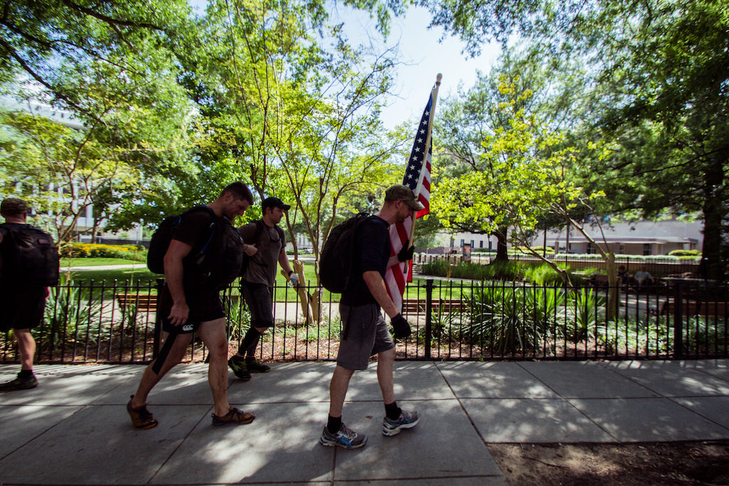Four people with backpacks walk on a sidewalk near trees, one carrying an American flag.