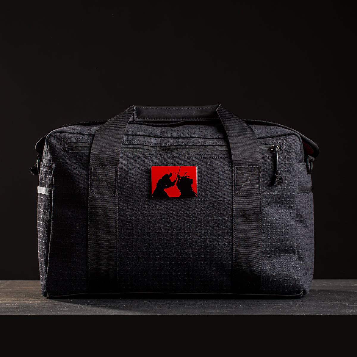 The Kit Bag x Carryology - GRXC2 Samurai 2.0 is a black duffel with a grid pattern, made from custom technical fabric, and features a red patch with two silhouettes—ideal for daily use or travel.