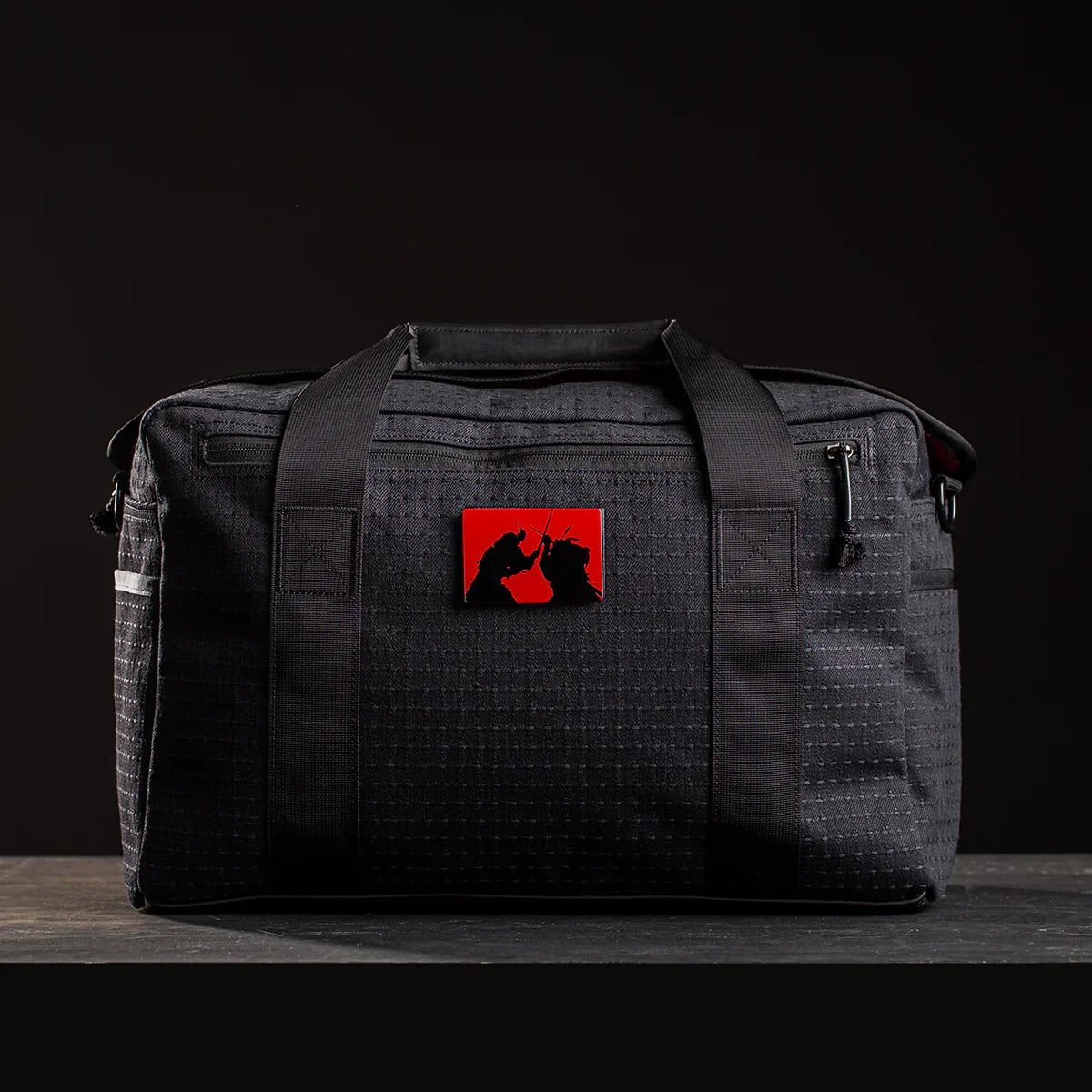 Black rucksack with dual handles and red patch showing samurai silhouettes, set on wooden surface