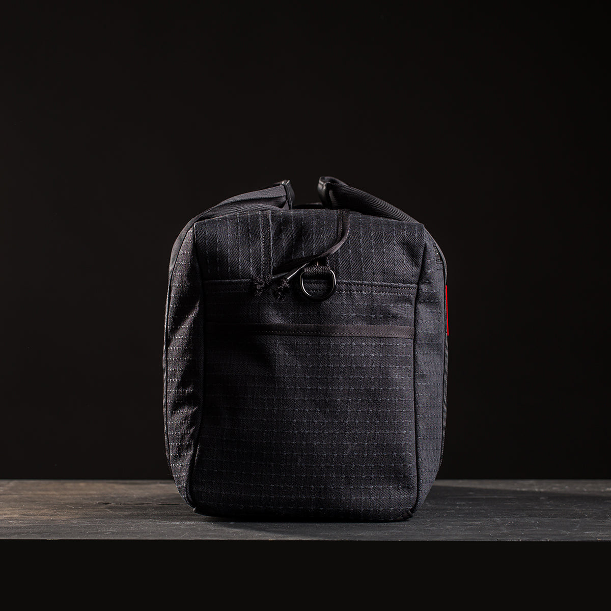The Kit Bag x Carryology - GRXC2 Samurai 2.0 is a black, rectangular bag made from Japanese denim with a zipper and front pocket, shown against a dark background.