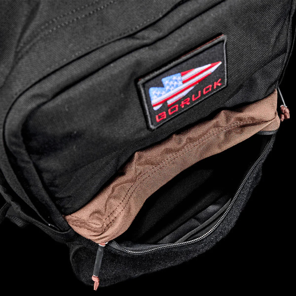 Close-up of black GORUCK backpack with American flag patch and partially open zipper showing brown interior
