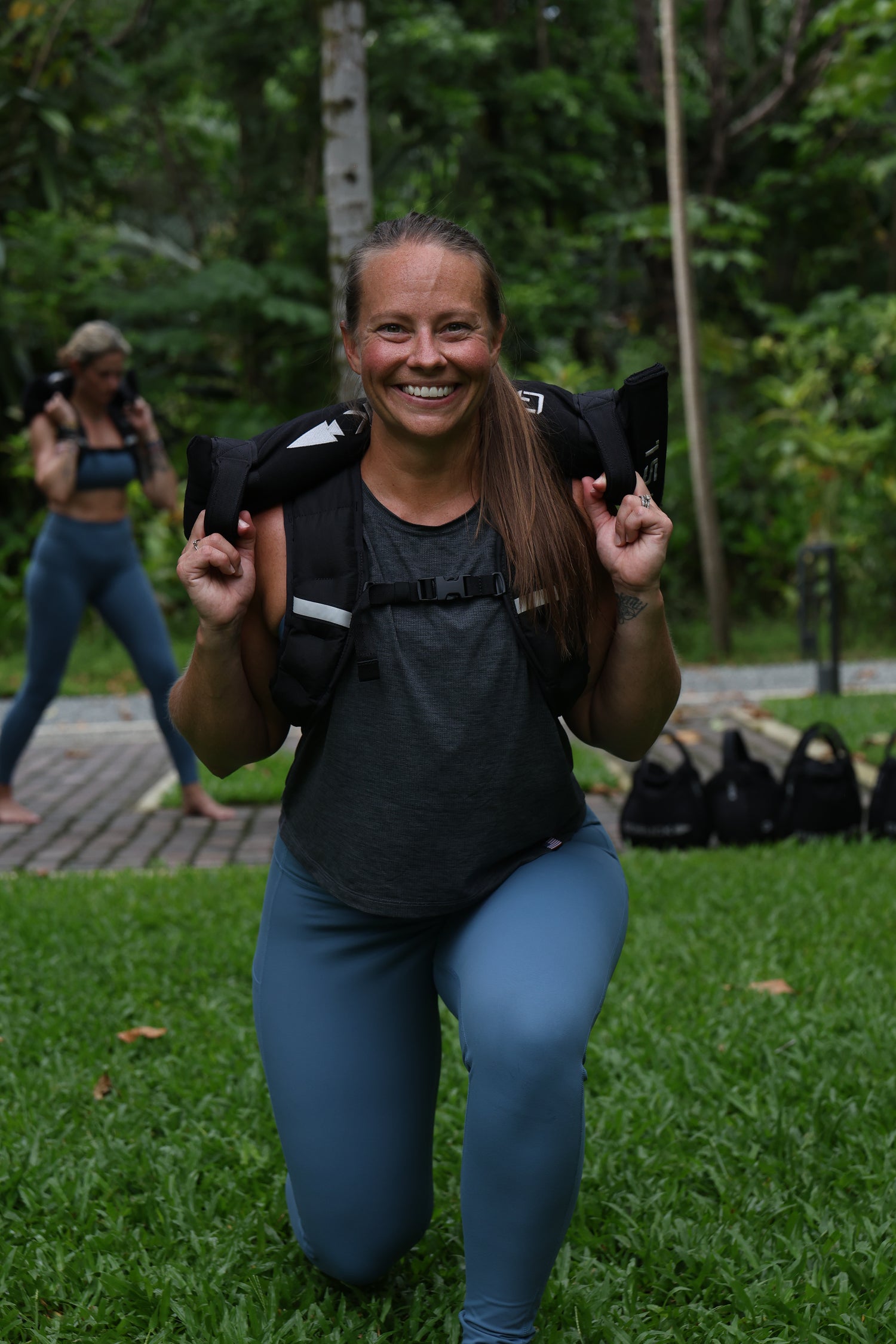 A woman smiles while doing a lunge outdoors, holding a weighted bag across her shoulders with the Patch - Ruck Fit Challenge to Start 2026 Strong.