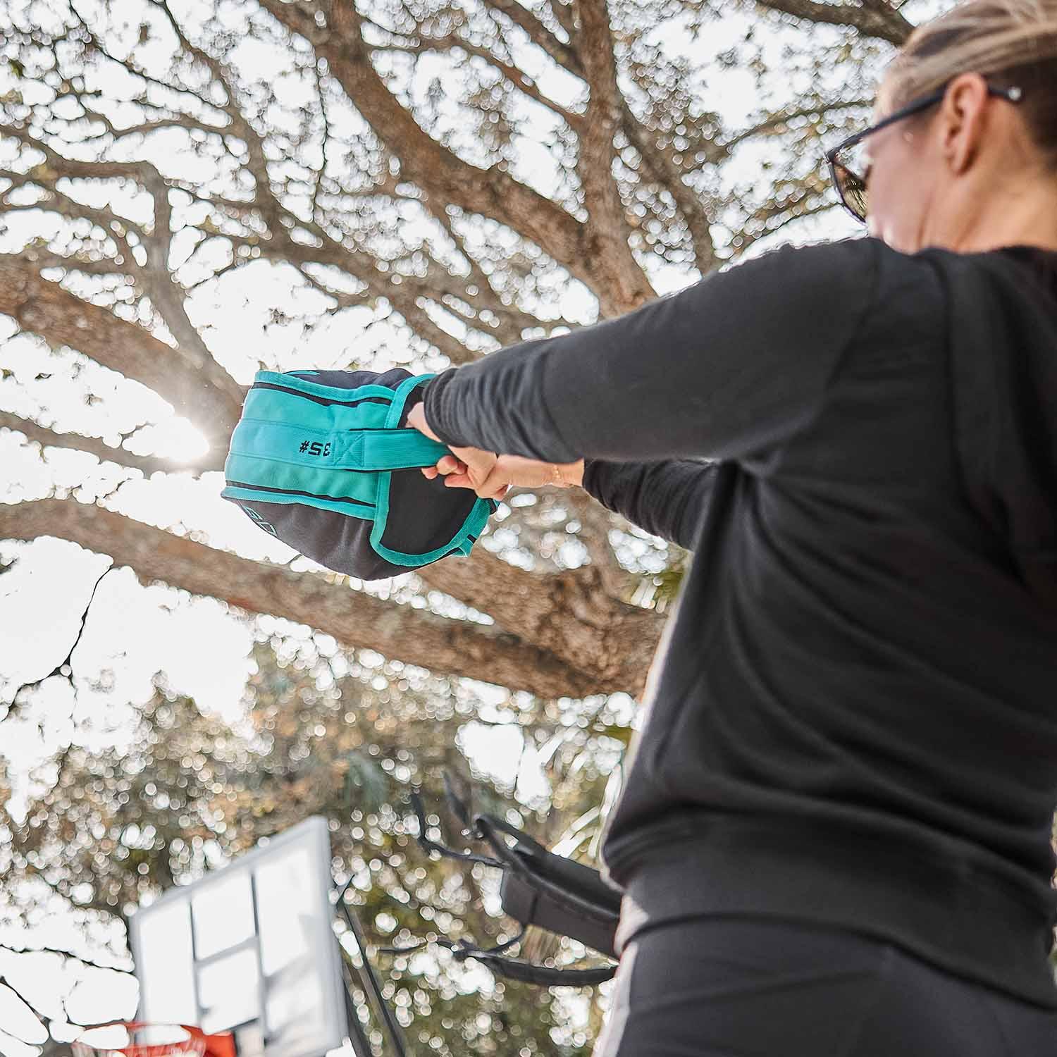 Person swinging teal and black 3.5 pound kettlebell outdoors with tree and basketball hoop background