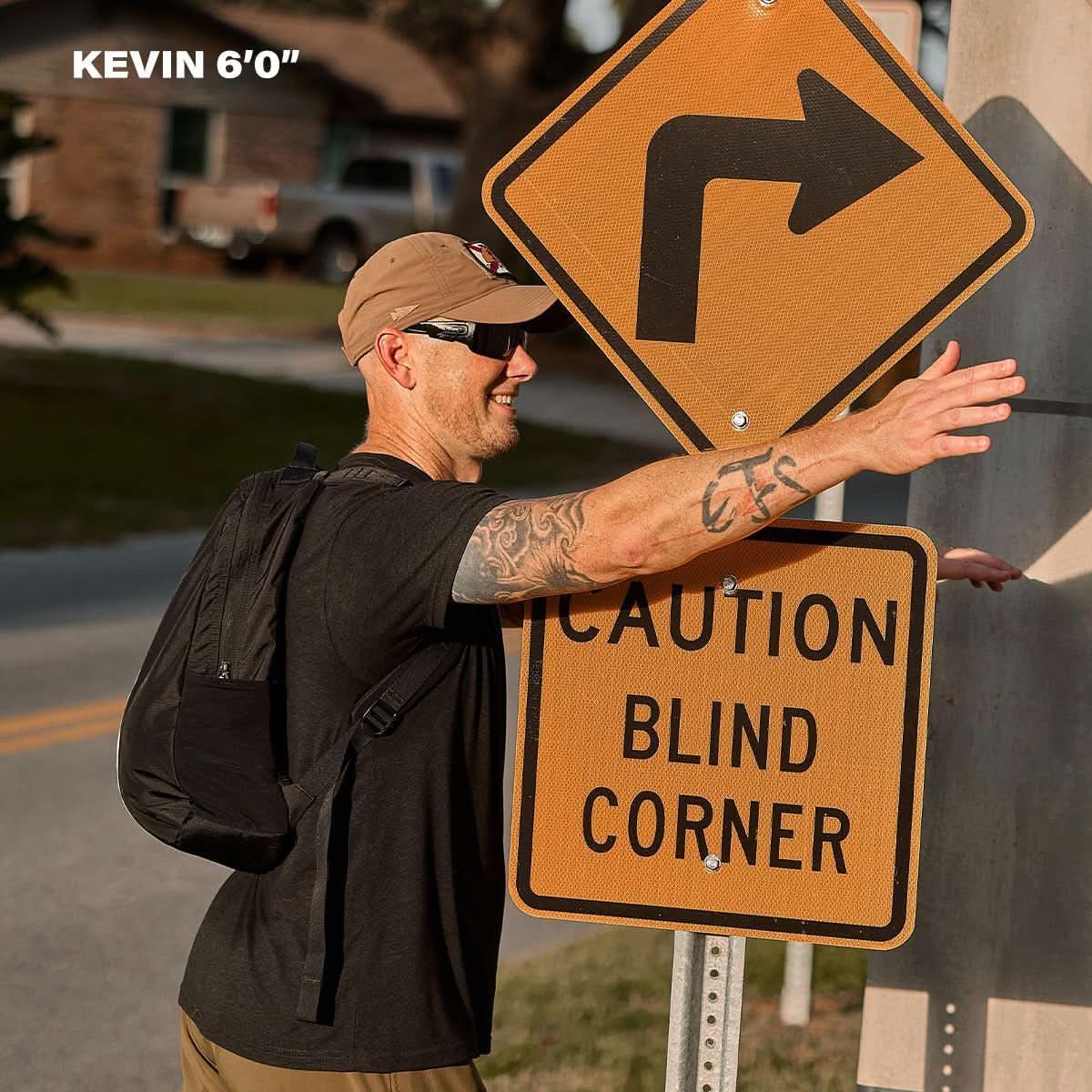 6-foot tall man wearing sunglasses, tan cap, and black backpack demonstrating arm reach next to a 'Caution Blind Corner' road sign