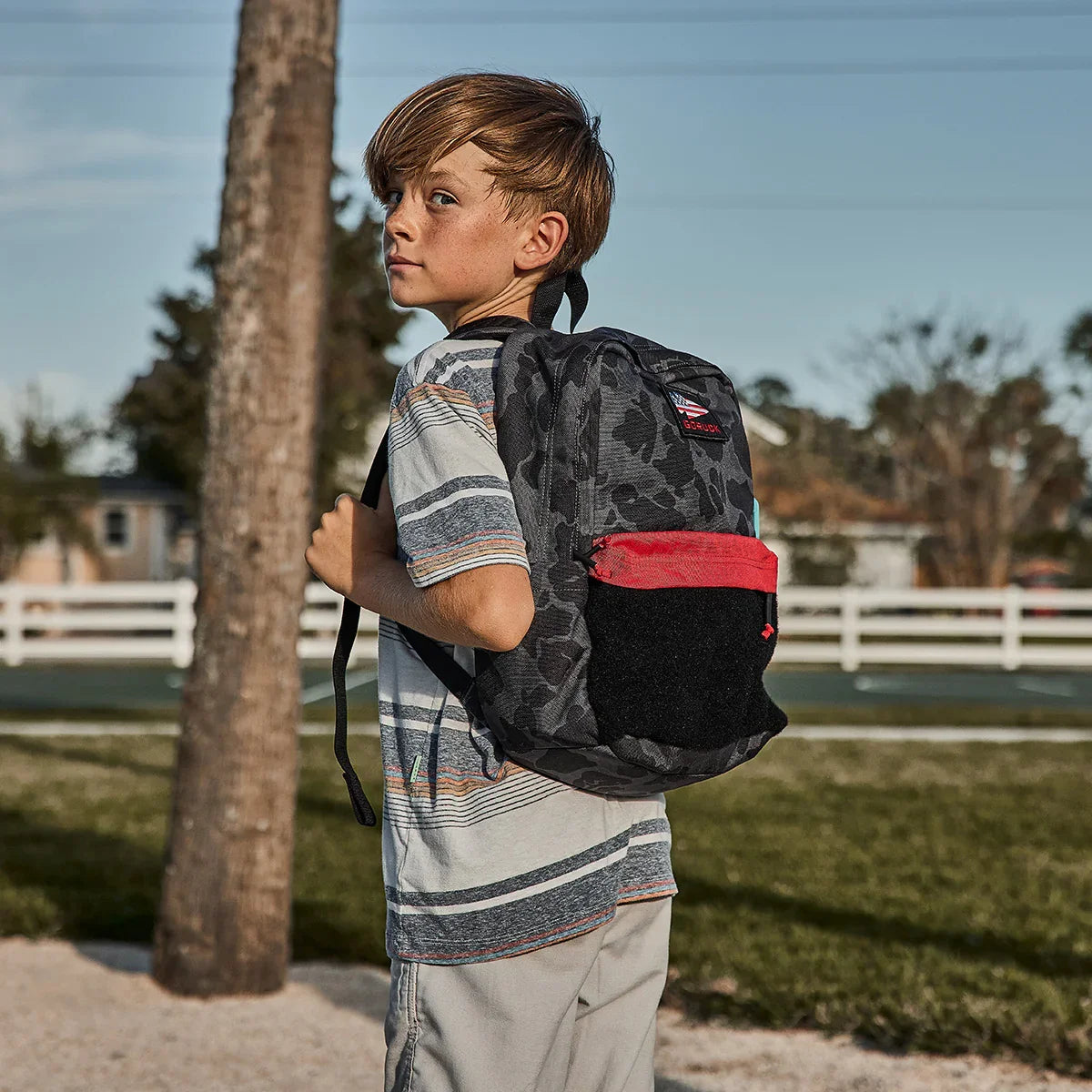 Young boy outdoors wearing a black camo and red GORUCK backpack with American flag patch
